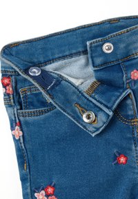 Jeans de denim azul con bordado floral en tonos de rosa y rojo. Presentan un cierre de botón y una tela texturizada. Detalles de costura visibles.