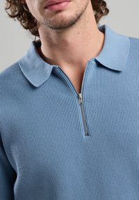 Light blue ribbed pullover with a zip collar. Features soft texture and a fitted shape, highlighting the zipper and collar design.
