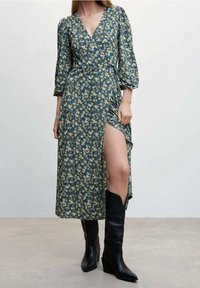 Blumen-Midi-Kleid in Dunkelgrün mit Puffärmeln, einem tiefen V-Ausschnitt und einem seitlichen Schlitz. Kombiniert mit schwarzen kniehohen Stiefeletten mit Absatz.