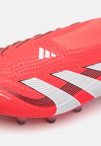 Chaussure de football rouge avec un dessus texturé, présentant des rayures blanches en diagonale, en matériau synthétique et des accents noirs. Semelle à crampons pour une meilleure traction.