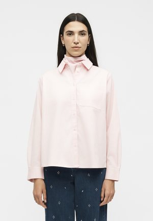 Žena s dlhými tmavými vlasmi, oblečená v svetloružovej košeli na gombíky cez zladený rolák a tmavomodré nohavice s malými bielymi vzormi.