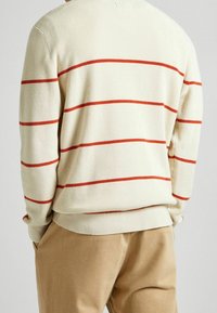 Cremefarbener, gerippter Pullover mit roten horizontalen Streifen, locker geschnitten und mit abgerundetem Saum, kombiniert mit beigefarbenen Hosen.