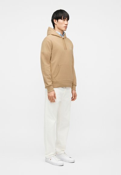 Νεαρός άνδρας που στέκεται, φοράει μπεζ φούτερ με κουκούλα, γαλάζιο πουκάμισο με γιακά, λευκό παντελόνι και λευκά αθλητικά παπούτσια, μπροστά σε σκέτο λευκό φόντο.