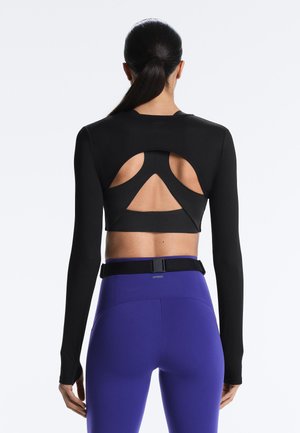 Ženska s črnim konjskim repom, oblečena v črno oprijeto majico z dolgimi rokavi s prerezi in visoko pasnimi vijoličastimi legicami s črnim pasom, stoji obrnjena stran.