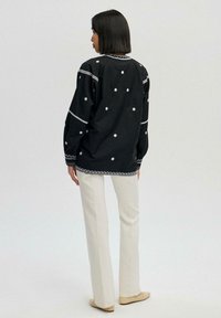 Black embroidered blouse with white floral patterns, long sleeves, and a round neckline, paired with cream-colored trousers and beige footwear.
