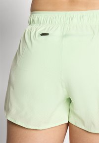 Close-up van lichtgroene korte broek met kleine perforaties en een achterzak met rits op de taille en de bovenbenen van een persoon.