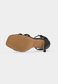 Μαύρο πέδιλο με λεπτά λουράκια και ψηλό τακούνι, σε προβολή από την πλευρά της καφέ σόλας, που δείχνει τετράγωνη μύτη και λεπτό τακούνι με λουράκι στον αστράγαλο με αγκράφα.