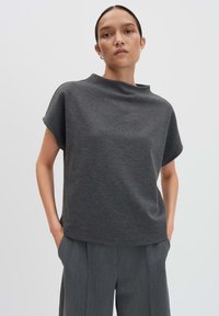 Vrouw in een antracietgrijze T-shirt met korte mouwen en bijpassende brede broek, staande met handen in de zakken tegen een eenvoudige achtergrond.