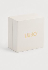 Kis, textúrázott, világos bézs színű, téglalap alakú doboz aranyozott "LIU·JO" márkanevével az elülső oldalán, sima háttér előtt.