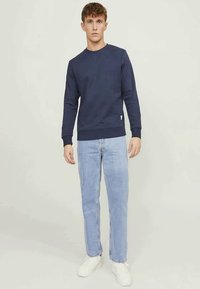Um jovem está de pé, de frente, vestindo um sweatshirt azul-marinho, jeans azul-claro e tênis brancos, contra um fundo claro e simples.