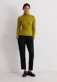 Olivgrüner, gerippter Pullover mit hohem Kragen, kombiniert mit schwarzen, taillierten Hosen und schwarzen Slippers. Einfaches, strukturiertes Strickdesign.