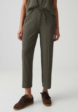 Person trägt olivgrüne strukturierte Hose mit Kordelzug in der Taille und braunen Schnürsneakern, steht auf einem hellgrauen Boden.