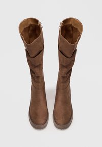 Brown slouchy knee-high boots made of suede with a soft texture, featuring a smooth interior and a rugged sole. Zipper detail at the back.