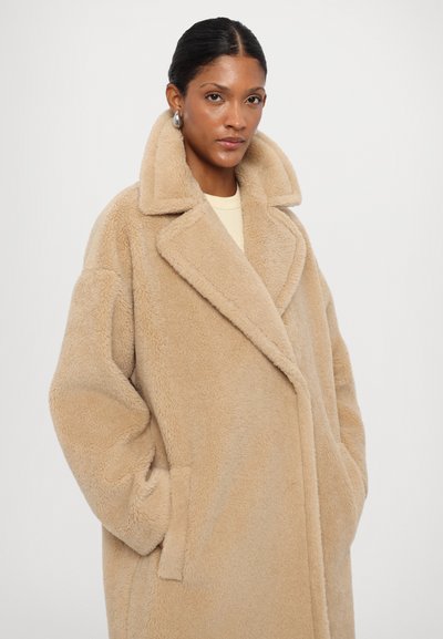 Beige oversized coat with a plush texture, wide lapels, side pockets, and a relaxed fit. The fabric appears soft and fluffy.