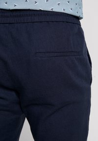 Detailní záběr na tmavě modré kalhoty s elastickým pasem a jednou hřebínkovou zadní kapsou pod světle modrou košilí s malými vzory plachetnic.