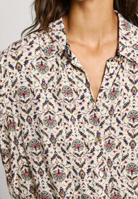 Skjorta med knappar i lätt tyg som har ett flerfärgat blommönster på en krämfärgad bakgrund; krage och knappdetaljer synliga.