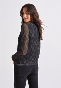 Zwarte doorschijnende top met luipaardprint, lange mouwen en een ontspannen pasvorm, gecombineerd met donkere jeans. Opvallende lichtgewicht stof en subtiele textuur.