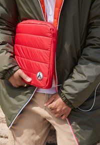 Custodia per laptop imbottita di rosso con motivo a righe orizzontali, con logo circolare JOTT. Tenuta vicino a una giacca verde con fodera arancione.