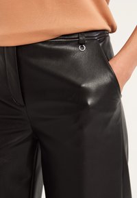 Pantalons en simili cuir noir avec une texture lisse, design à jambe large, poche avant et accent en métal argenté sur la taille.