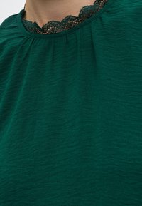 Gros plan d'un haut en tissu texturé vert foncé avec un bord en dentelle noire visible au niveau du décolleté sur la peau.