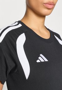 Μαύρο αθλητικό μπλουζάκι με λευκές λεπτομέ ρειες, με κοντά μανίκια και στρογγυλή λαιμόκοψη. Δύο λευκές ρίγες διατρέχουν τους ώμους. Εμφανίζεται το λογότυπο adidas.