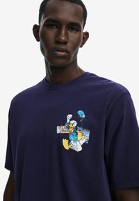 Tmavě modré bavlněné tričko s barevným potiskem Káčera Donalda na hrudi, který zobrazuje vztyčenou pěst a různé vzory.