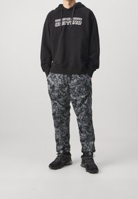 Černá mikina s kapucí s bílým logem, doplněná o černé tepláky s potiskem a šedým textem. Model má na sobě černé sportovní boty.