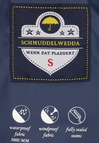 Tela navy con una etiqueta textil que muestra "Schmuddelwedda", con un logo de paraguas, costuras amarillas y símbolos de impermeable y a prueba de viento.