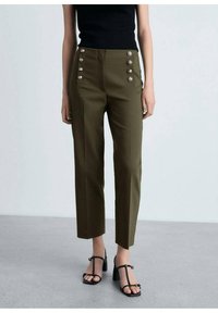 Vrouw in olijfgroene high-waisted broek met gouden knopen en zwarte T-strap sandalen met hakken, staand op een grijze vloer.