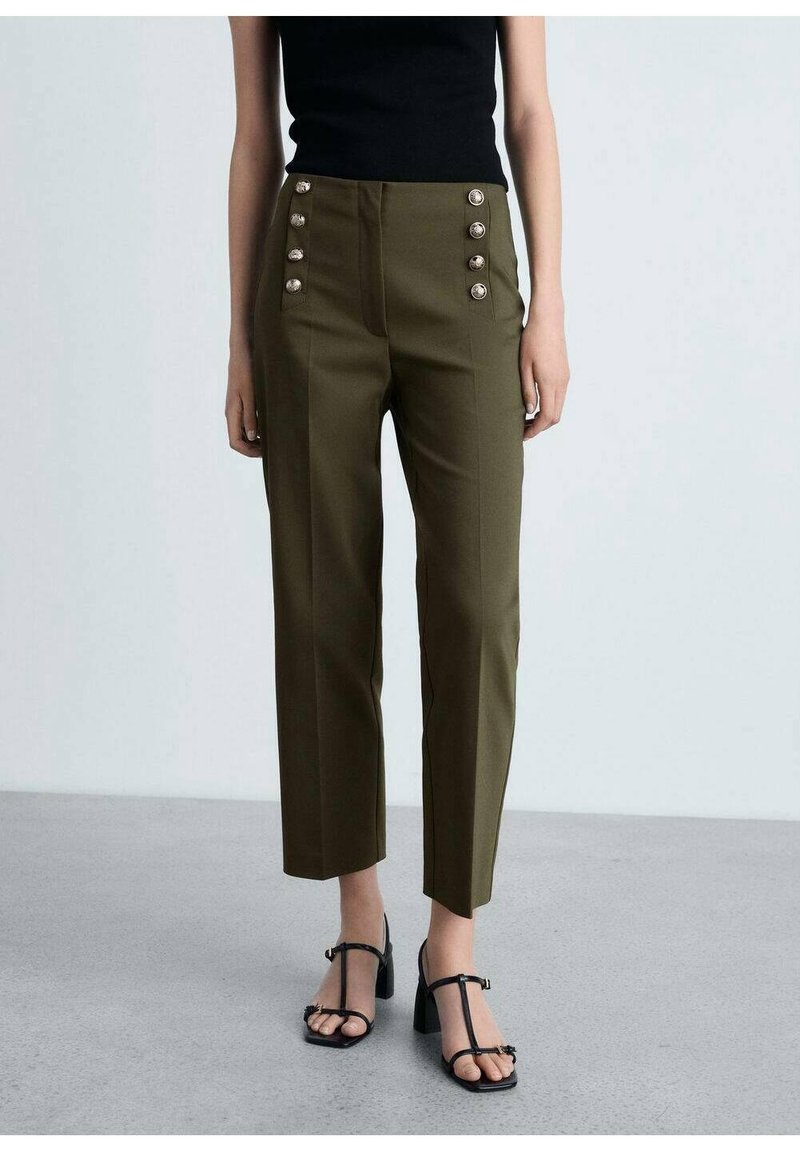 Vrouw in olijfgroene high-waisted broek met gouden knopen en zwarte T-strap sandalen met hakken, staand op een grijze vloer.