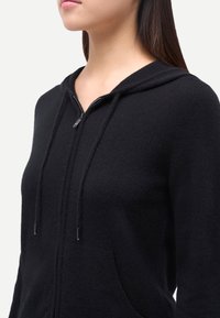 Svart zip-up hoodie i mjukt tyg, med en känguruficka, justerbar dragsko i huvan och silvriga detaljer i metall.