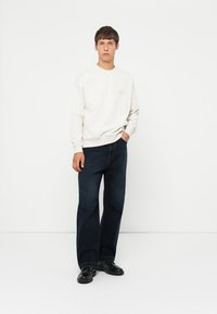 Sudadera beige claro con cuello redondo, combinada con jeans rectos de mezclilla oscura. El modelo está de pie sobre un fondo liso, con zapatos negros.