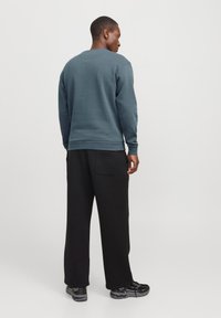 Hombre de pie de espaldas, vistiendo un sudadera azul, pantalones negros holgados y zapatillas deportivas negras, contra un fondo blanco liso.