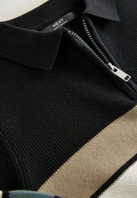 Mustapohjainen, vetoketjullinen polo, jossa on teksturoitu kangas. Mukana on yksivärinen musta sävy, kontrastiväri beige raita ja hopeanvärinen vetoketju.