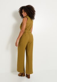 Combinaison vert olive avec une taille ceinturée, sans manches et jambes larges. Le tissu a une finition texturée et des poches latérales. Talons compensés marron.