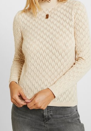 Vrouw in een beige gebreide trui met patroon en lange mouwen en grijze spijkerbroek, die de zoom van de trui met haar handen aanpast.