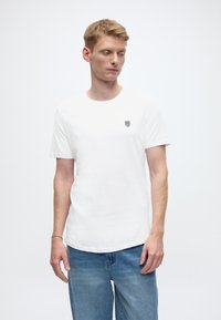 Bijela pamučna majica sa zaobljenim izrezom i kratkim rukavima. Na prsima se nalazi mali crni logo, u kombinaciji sa svijetloplavim trapericama.