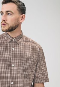 Camisa de manga corta a cuadros marrones con cuello abotonado, bolsillo frontal y botones blancos, fabricada con tela ligera.