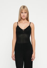 O tânără cu părul blond drept poartă un top negru fără mâneci cu bretele subțiri și pantaloni negri asortați, stând în fața unui fundal alb.