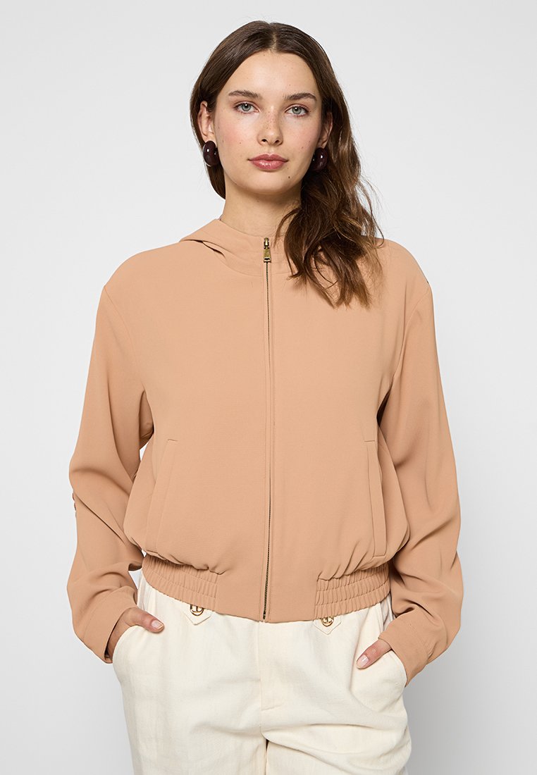Chaqueta con cremallera de color melocotón, con capucha, cintura elástica, bolsillos laterales y mangas largas. El tejido es suave, ligero y fluido.
