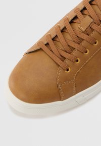 Brown leather sneaker featuring round toe, textured surface, and lacing with gold eyelets. White rubber sole with subtle texture along edges.