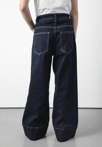 Szerokie dżinsowe spodnie w ciemnym indygo z białymi szwami, z wysokim stanem i dwiema tylnymi kieszeniami z zaostrzonym klapkiem.