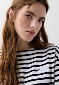 Striped long-sleeve top in white and navy, featuring a blue logo patch on the left chest, made of soft fabric with a relaxed fit.