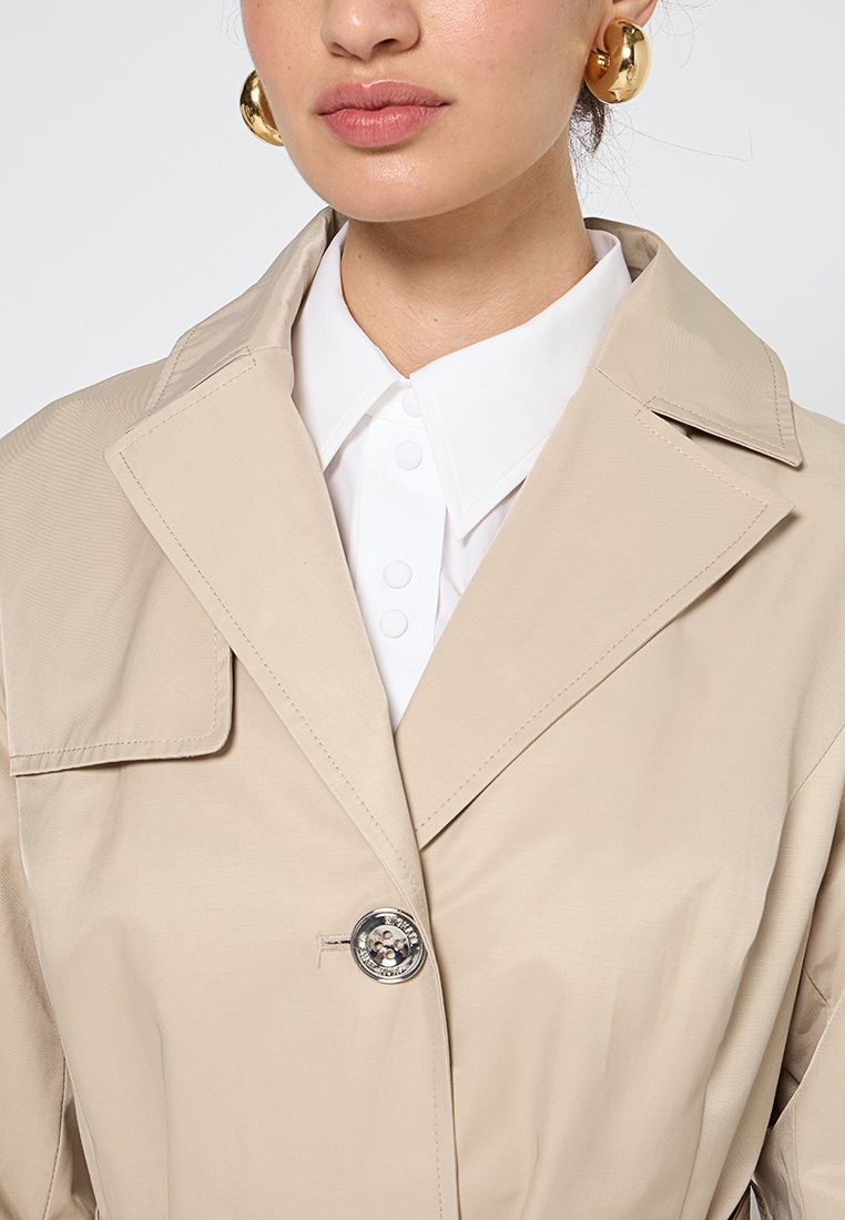 Manteau beige avec un col structuré, fermeture à boutons et texture subtile. Porté par-dessus une chemise blanche avec des boutons proéminents.