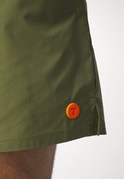 Shorts verts en tissu léger avec un logo circulaire orange arborant le numéro 7 sur l'ourlet inférieur, texture lisse et lignes épurées.