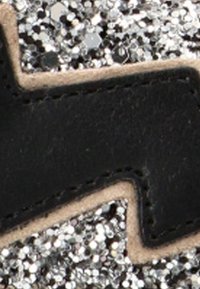 Rayo negro cosido sobre un fondo de brillantina plateada con un borde beige alrededor del rayo.