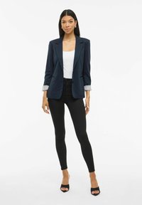 O femeie stă îmbrăcată într-un sacou bleumarin cu manșete desfăcute, un top alb, pantaloni skinny negri și sandale negre cu vârful deschis, pe un fundal alb simplu.