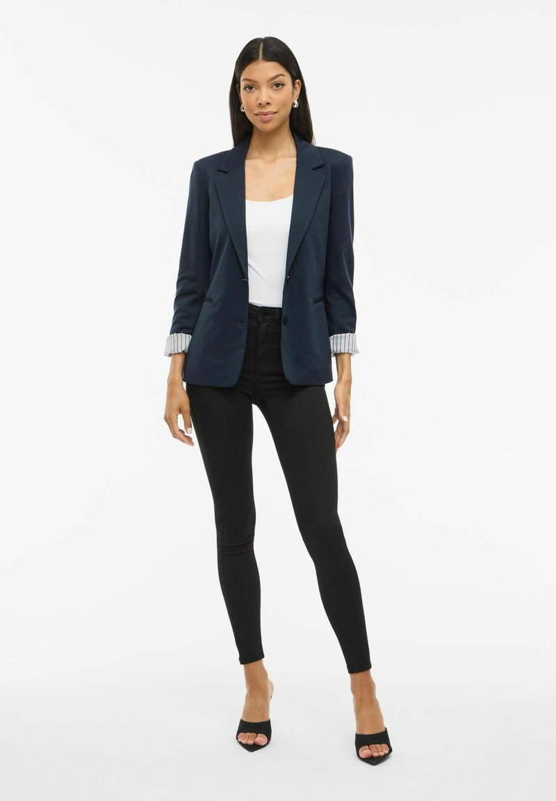 O femeie stă îmbrăcată într-un sacou bleumarin cu manșete desfăcute, un top alb, pantaloni skinny negri și sandale negre cu vârful deschis, pe un fundal alb simplu.
