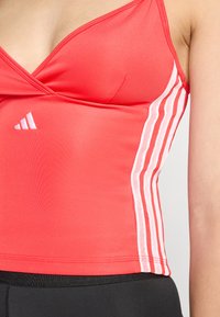 Crvena atletska crop majica s dekolteom u obliku slova V, s bijelim mrežastim prugama i tri bijele adidas pruge sa strane. Glatka tekstura tkanine.