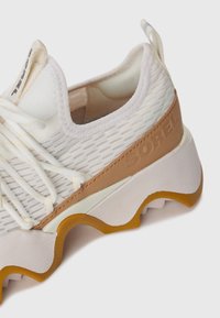 Hvit og beige mesh treningsko med bølgete såle og "SOREL"-logo preget på siden av hælen.
